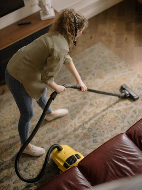 A woman with curly hair, wearing a green shirt and dark jeans, is performing surface cleaning using a yellow vacuum cleaner with a hose attachment on a patterned area rug in a living room. The room fe