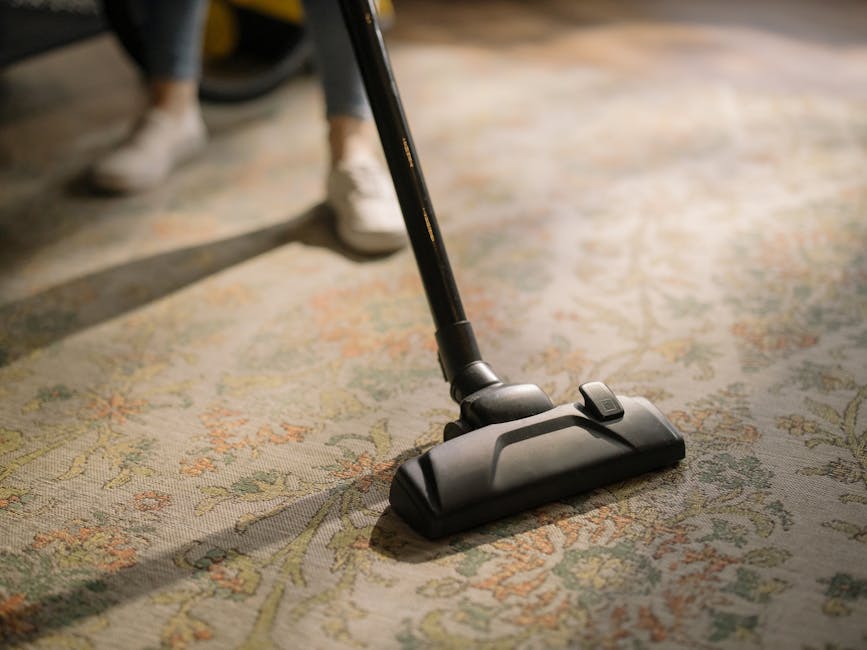 A woman with curly hair, wearing a green shirt and dark jeans, is performing surface cleaning using a yellow vacuum cleaner with a hose attachment on a patterned area rug in a living room. The room fe